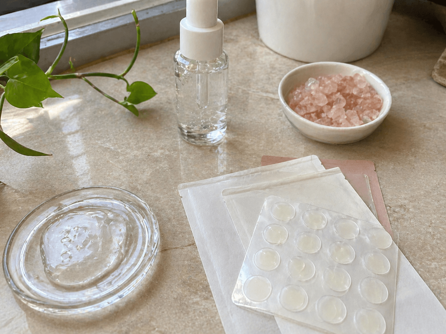 A handwritten ingredient checklist surrounded by petri dishes, a serum dropper, pink Himalayan salt, and a sheet of hydrocolloid patches on a soft beige surface